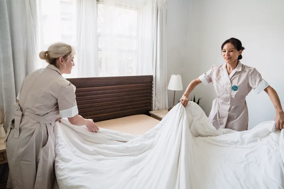Zwei Reinigungskräfte beziehen in einem Schlafzimmer gemeinsam ein frisches Bettlaken